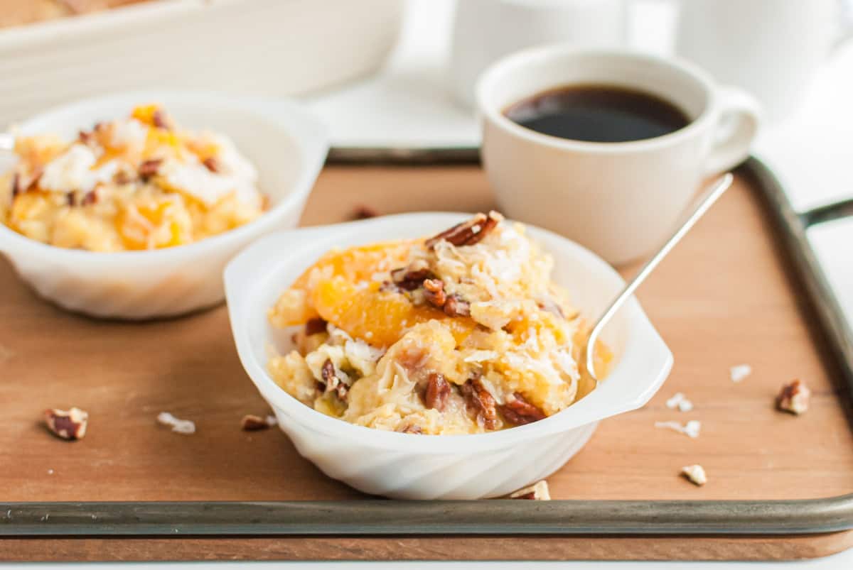 Milk glass dish with dollop of peach dump cake and spoon. On a wooden serving tray with mug of coffee.