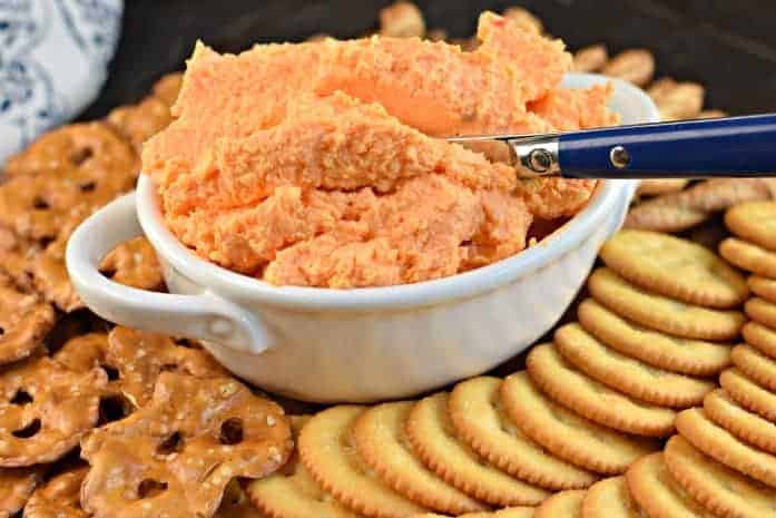 White bowl with pimento cheese spread and a blue knife. Tray with ritz crackers and pretzels for serving.