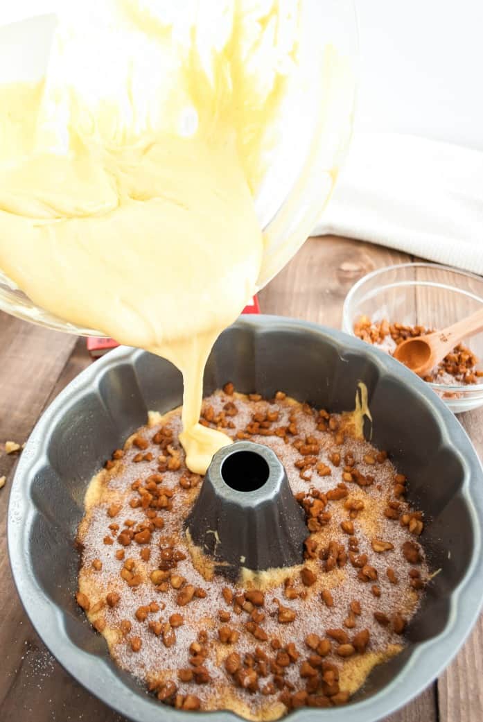 Bundt pan with half the cake batter, nuts, cinnamon sugar, and a glass bowl pouring the remaining cake batter over the top.