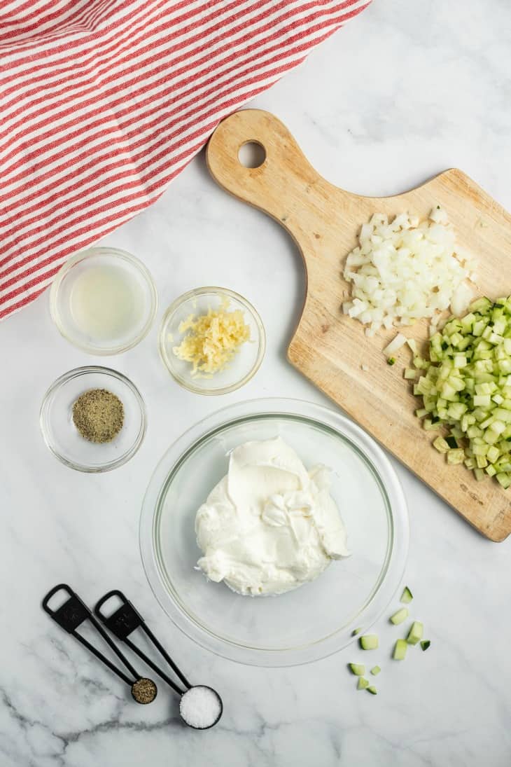 Ingredients needed for tzatziki on marble counter, including greek yogurt, cucumber, onion, seasonings, and lemon juice.