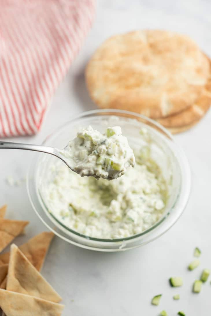 Clear glass bowl of tzatziki cucumber sauce with a metal spoon lifting a scoop out of the bowl.