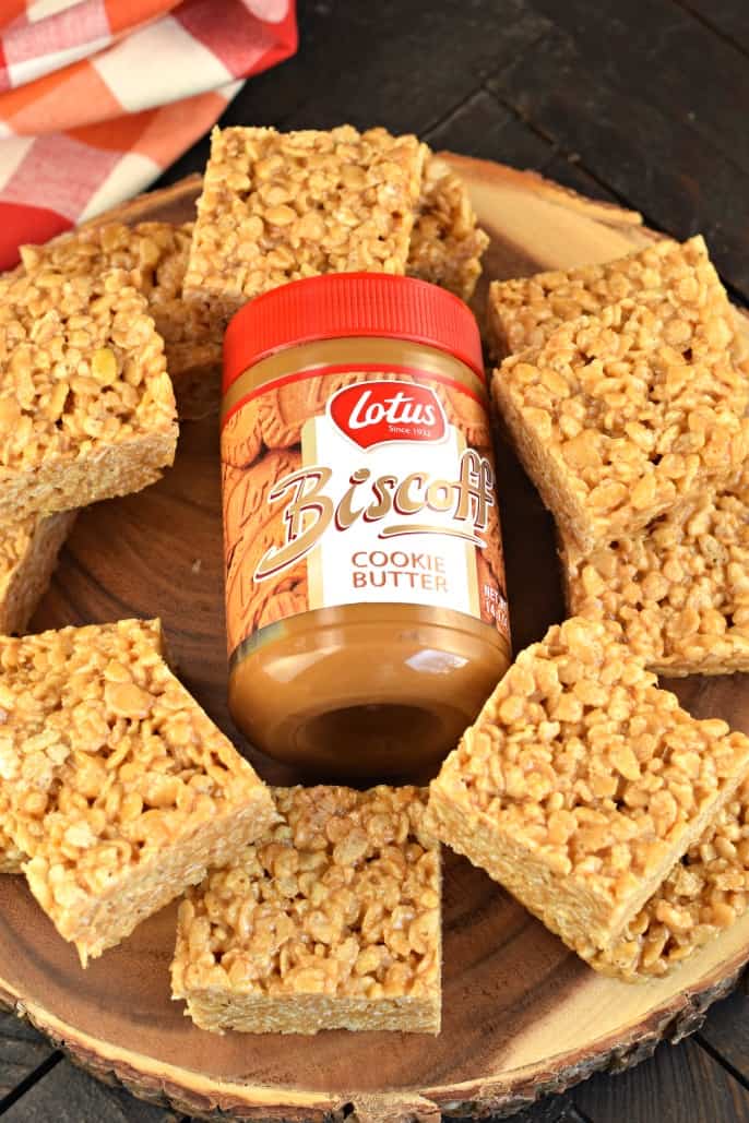 Biscoff rice krispie treats on a wooden cake plate with a jar of cookie butter on the plate too.