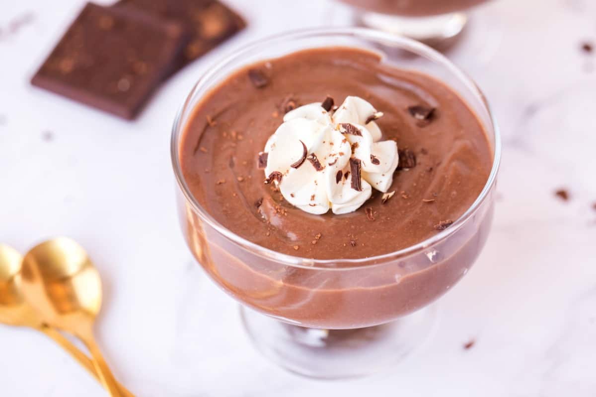 Clear glass dessert dish with chocolate pudding. Topped with whipped cream, chocolate shavings, and a gold spoon on the side.