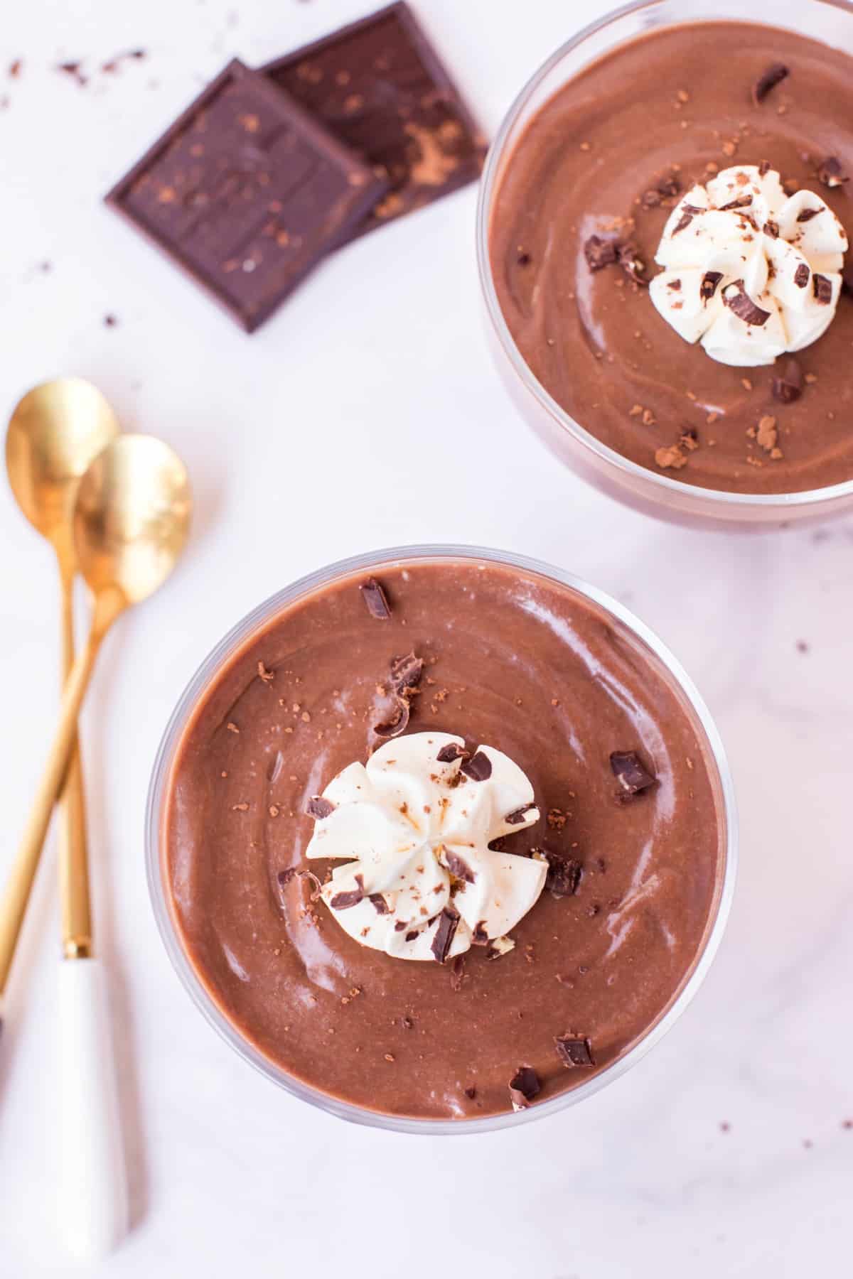 Two clear glass bowls of chocolate pudding with whipped cream. Two gold spoons on side of bowls.