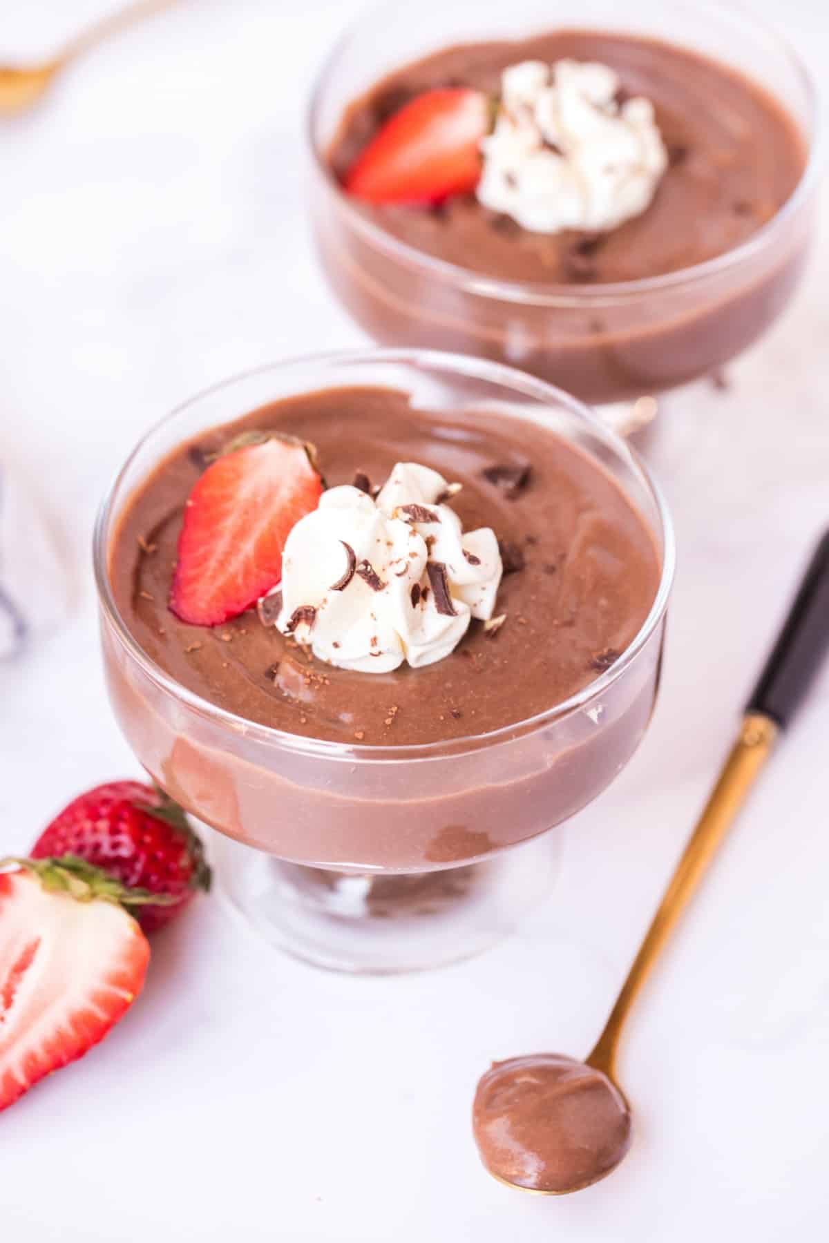 Two glass dishes with chocolate pudding, whipped cream, and strawberries. Spoonful of chocolate pudding on side.