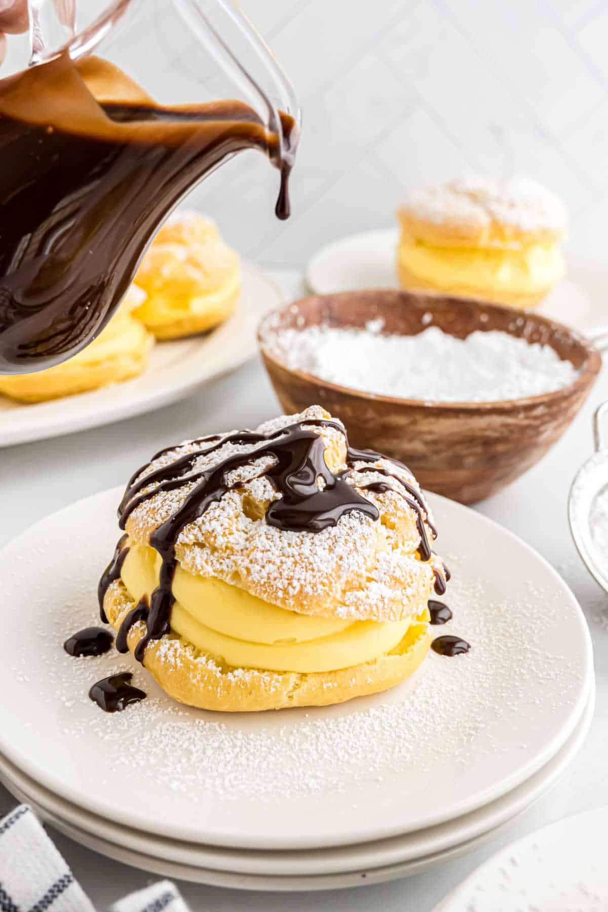 A cream puff topped with powdered sugar is elegantly drizzled with chocolate sauce from a glass pitcher, creating a decadent treat that highlights the delicate texture of the cream puff.