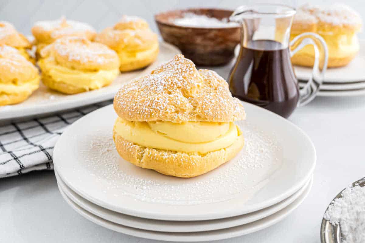 A delicious cream puff filled with yellow custard sits on a white plate, dusted with powdered sugar.