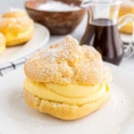 Cream puff filled with vanilla custard, dusted with powdered sugar, on a white plate with syrup in the background—a delightful cream puff dessert for any occasion.