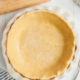 Homemade pie crust in a pie plate before baking with a rolling pin in background.