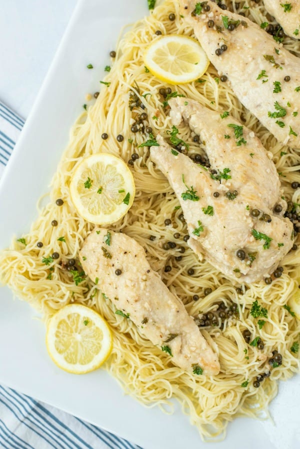 Chicken piccata over pasta on a large white serving platter, garnished with capers and lemon slices.