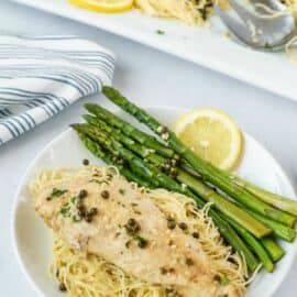 Chicken piccata on a white dinner plate with pasta noodles, grilled asparagus, and a slice of lemon.