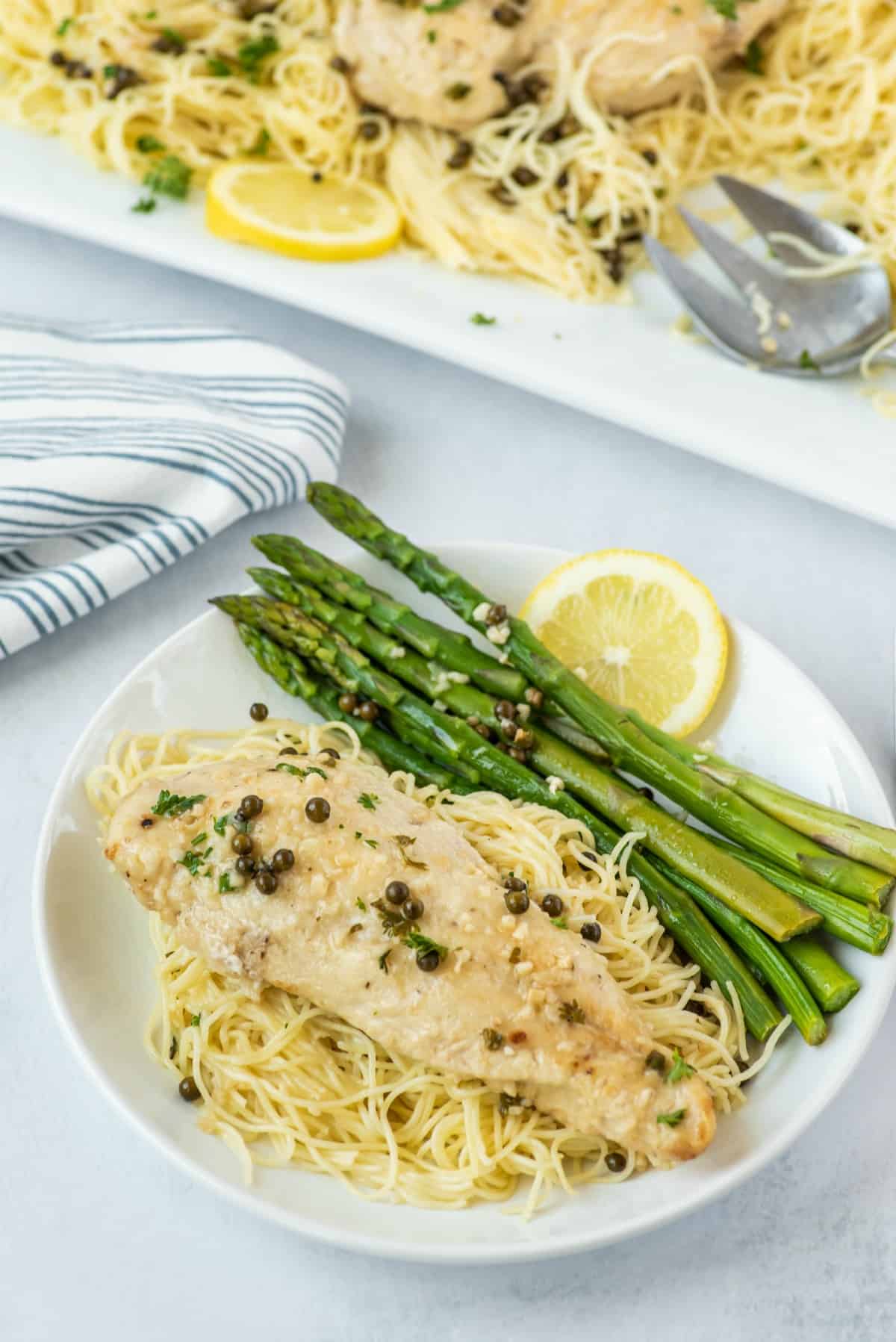 Chicken piccata on a white dinner plate with pasta noodles, grilled asparagus, and a slice of lemon.