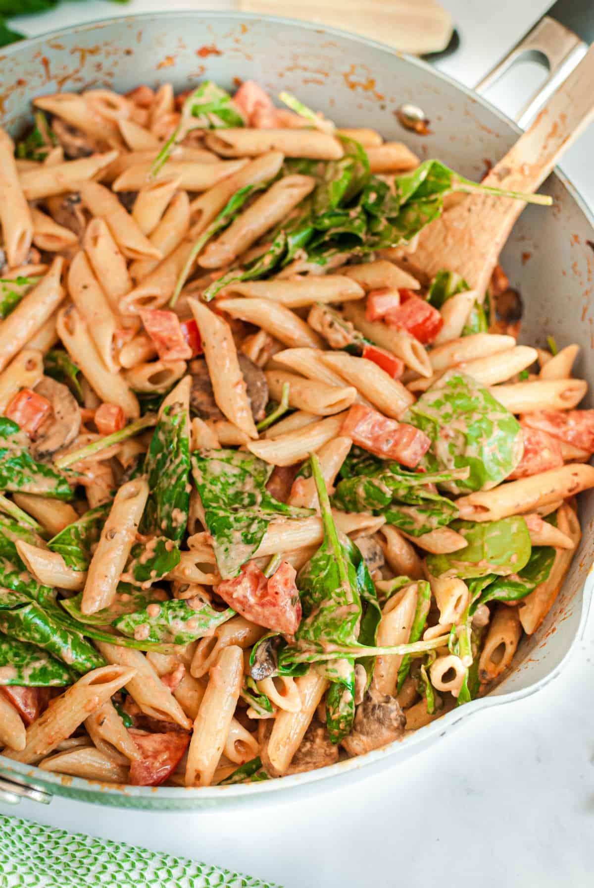 Large white skillet filled with cooked pasta in cream sauce with mushroom, tomatoes, and spinach.