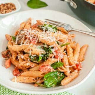 Penne pasta in a white bowl with tomato cream sauce, spinach, tomatoes, and mushrooms.