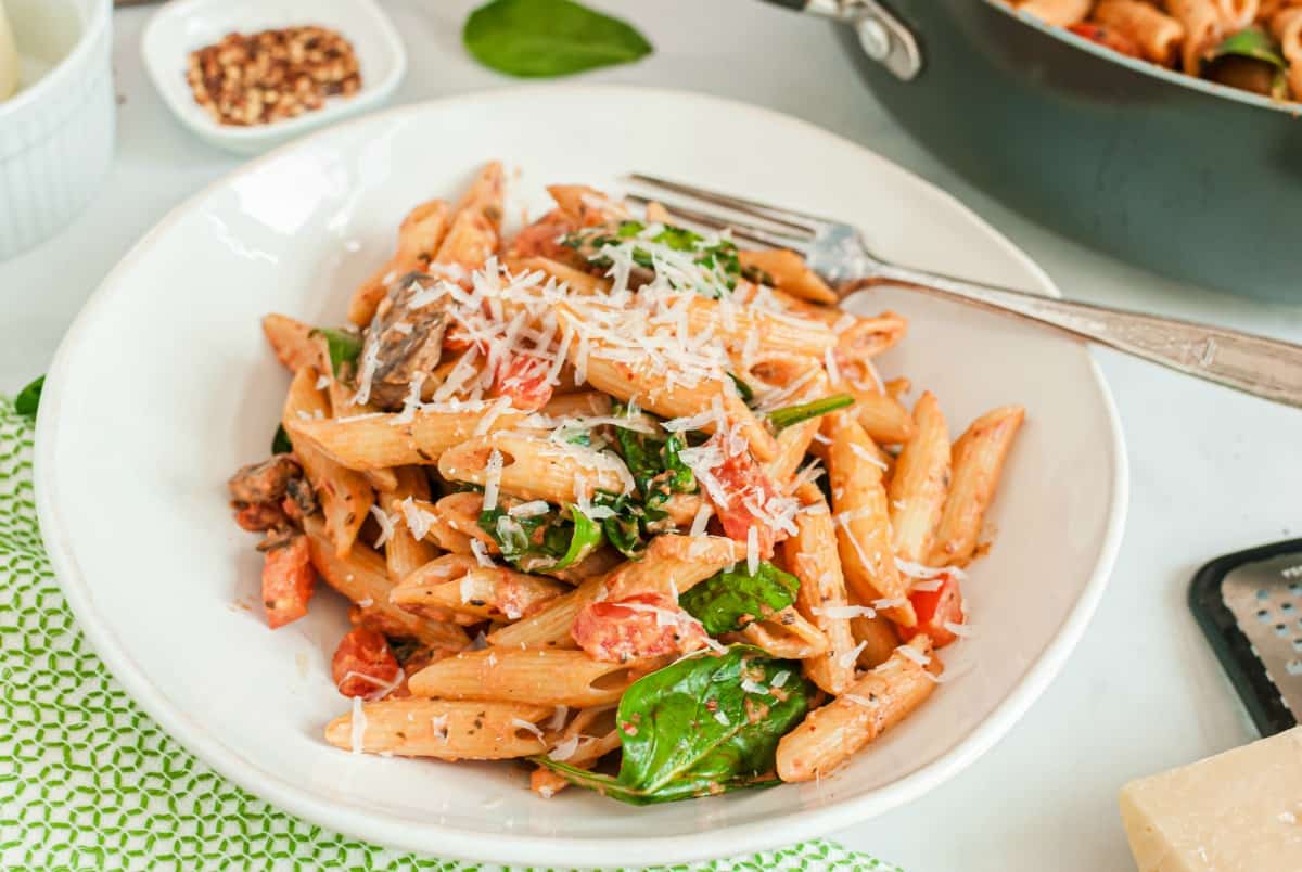 Penne pasta in a white bowl with tomato cream sauce, spinach, tomatoes, and mushrooms.