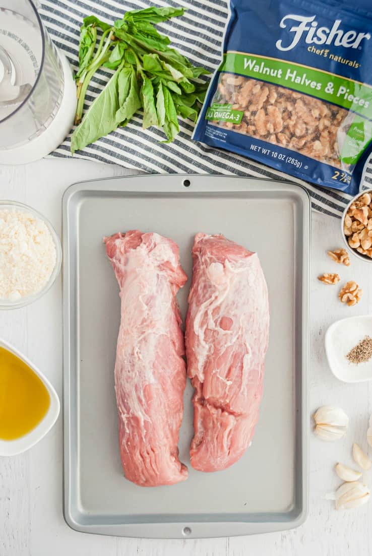 Two raw pork tenderloins on a baking sheet with a bag of walnuts and basil on a wire rack in the background.
