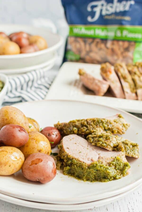 White dinner plate with sliced pesto topped pork tenderloin and baby potatoes, with a bag of Fisher Nuts walnuts in the background.