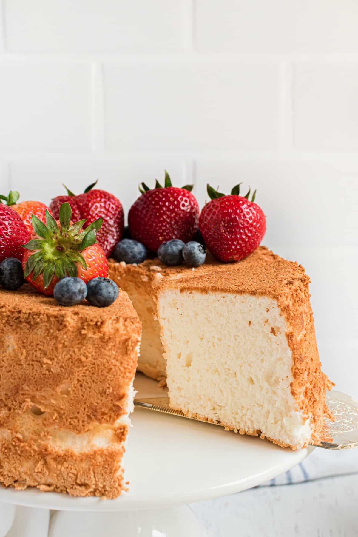 Angel food cake on a white cake platter topped with fresh berries. One slice removed.