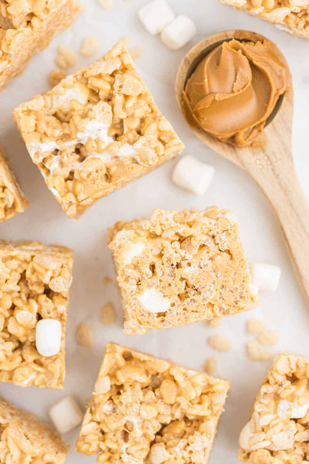 Biscoff Rice Krispie Treats with visible marshmallows, next to a wooden spoon of peanut butter on a white surface.
