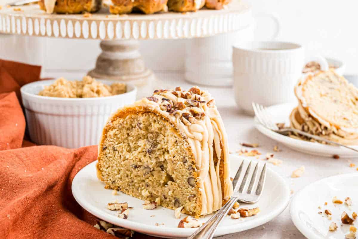 Slice of brown sugar pound cake with icing and chopped pecans on a plate, with a fork and cake stand in the background.