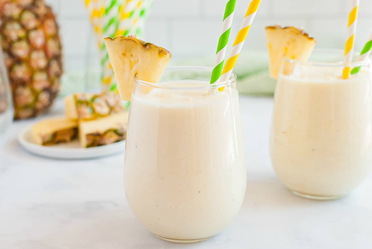 Dole whip in two stemless wineglasses garnished with fresh pineapple slices. Served with yellow and green striped paper straws.