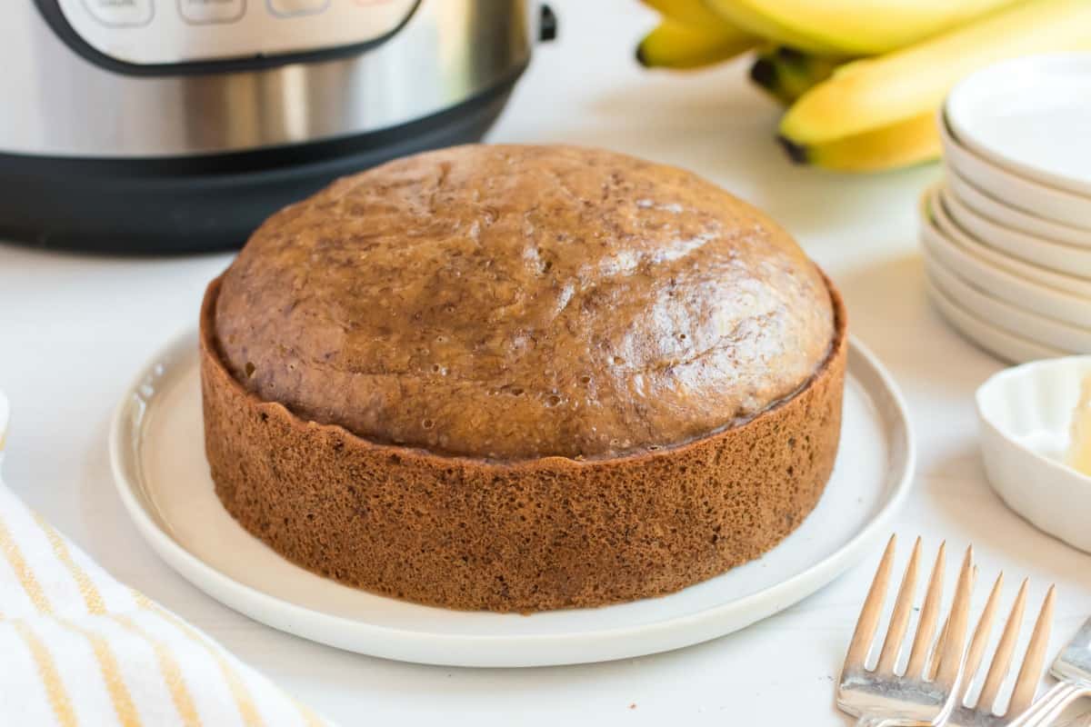Banana bread on white serving platter with stack of plates, bananas, and instant pot in background.