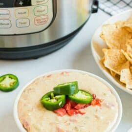 Queso in a white bowl with instant pot in background. Garnished with rotel tomatoes and jalapenos.