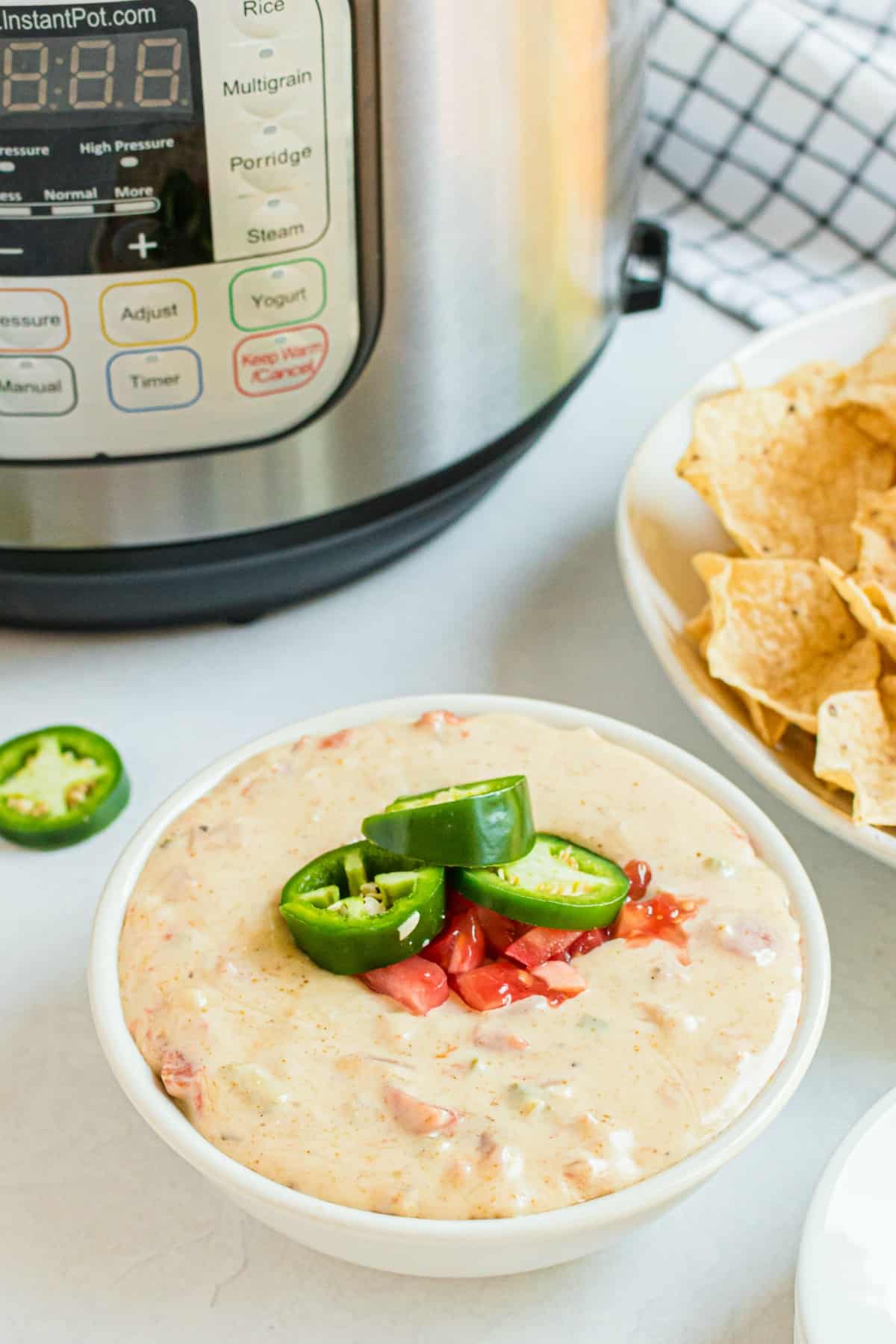 Queso in a white bowl with instant pot in background. Garnished with rotel tomatoes and jalapenos.