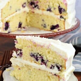Slice of layered lemon blueberry cake on a white plate, with whole cake on cake stand in background.