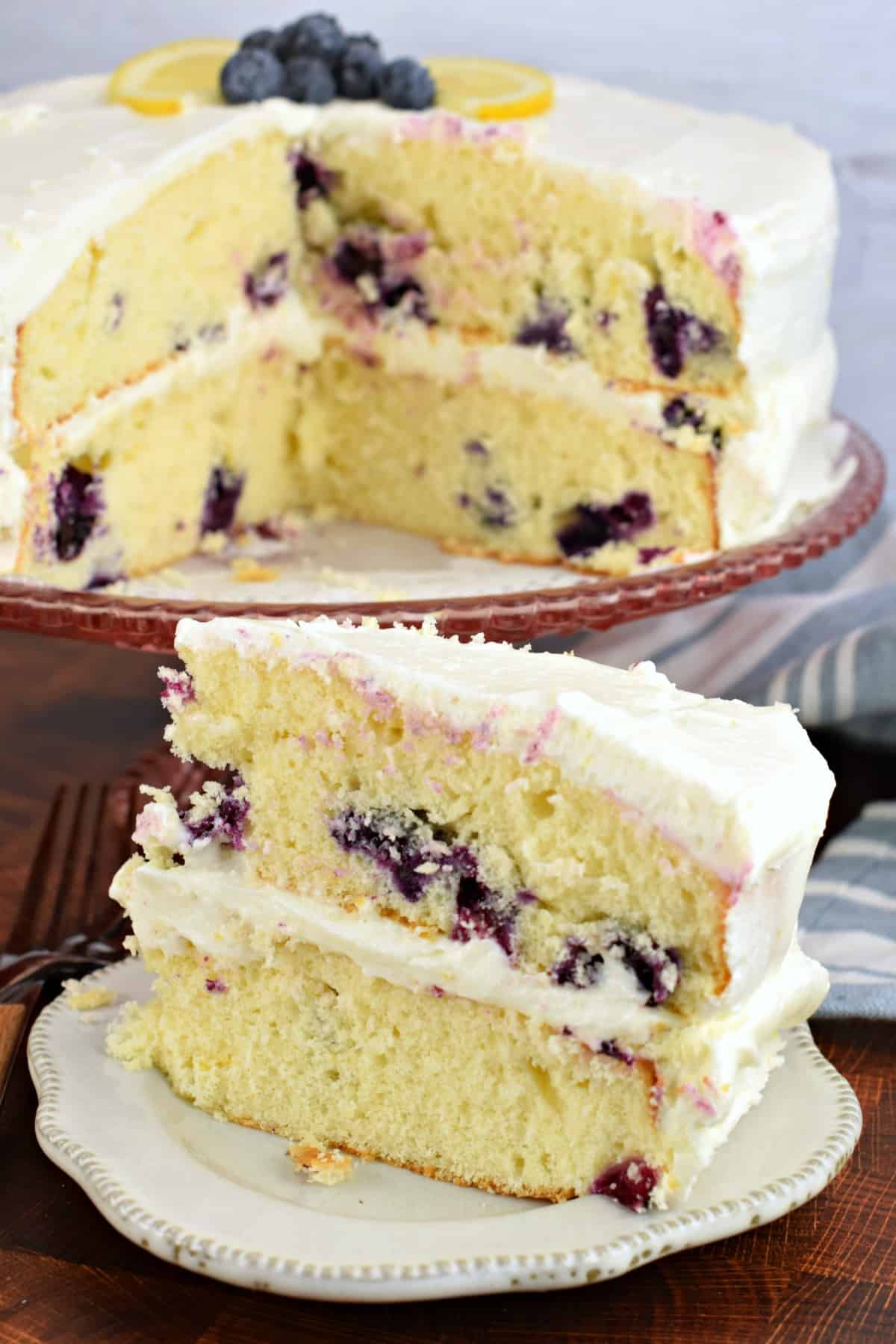Slice of layered lemon blueberry cake on a white plate, with whole cake on cake stand in background.