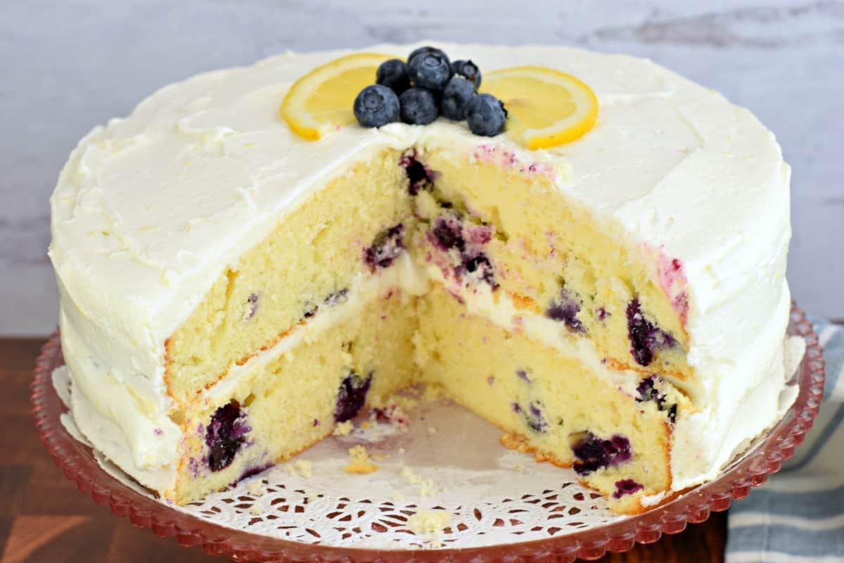 Lemon Blueberry cake on a wooden cake stand with white doily. Slices removed so you can see the center.