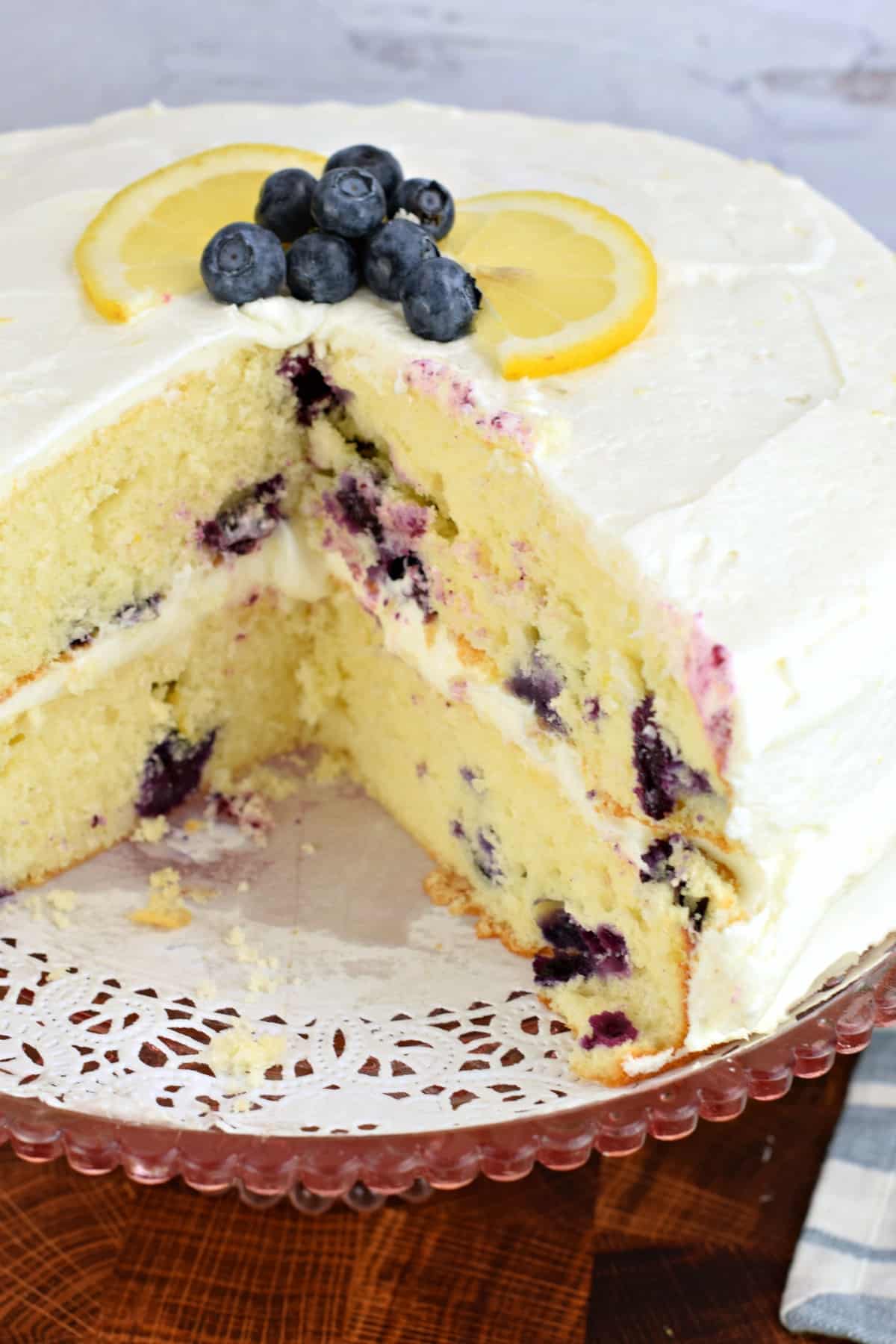 Lemon cake with blueberries on a doily topped wooden cake stand, with a slice removed. Garnished with fresh blueberries and slices of lemon.