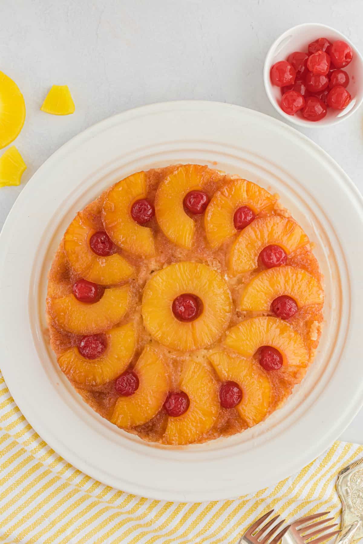 Pineapple upside down cake on a white cake platter with a small bowl of cherries in background.