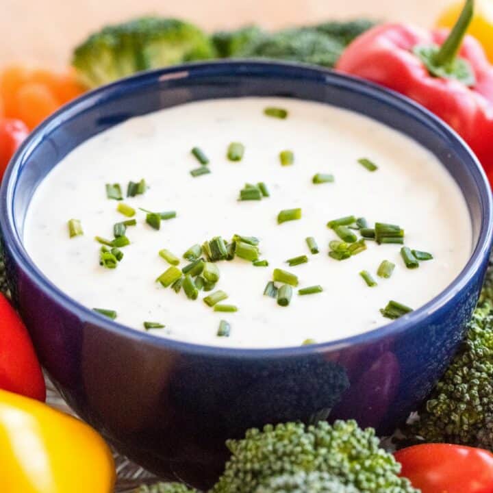 Dark blue bowl filled with homemade ranch dressing, topped with chives, and surrounded by fresh cut vegetables.