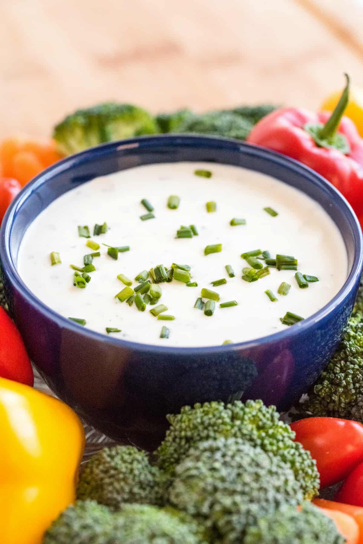 Dark blue bowl filled with homemade ranch dressing, topped with chives, and surrounded by fresh cut vegetables.