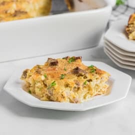 A slice of breakfast casserole with sausage and cheese sits on a white plate, while more breakfast casserole awaits in the background.