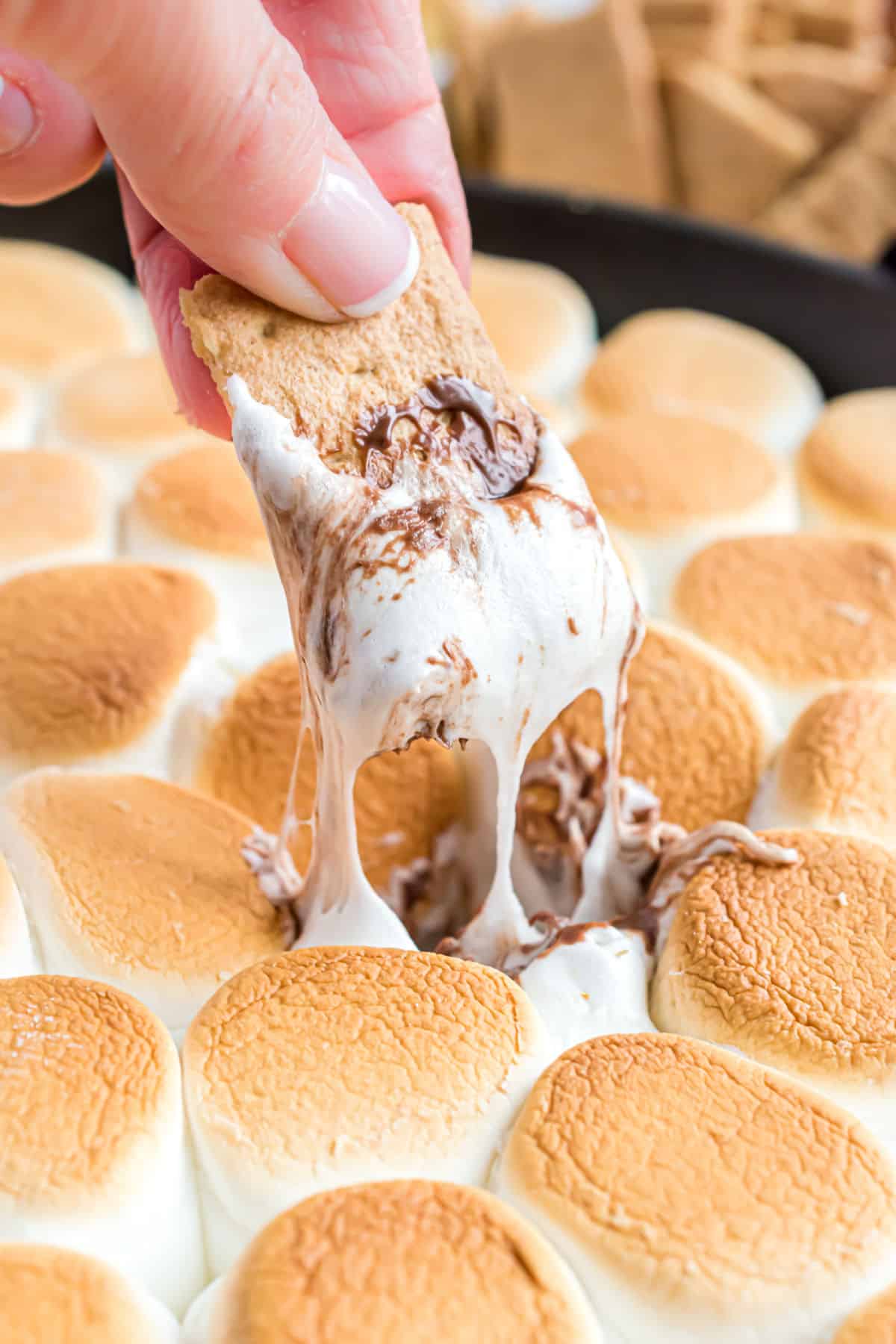 Graham cracker being dipped into a skillet of chocolate and melted marshmallow.