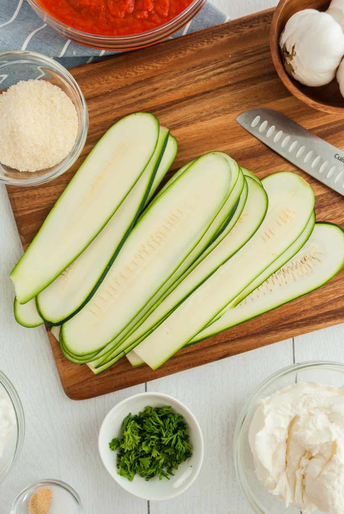Thinly sliced zucchini strips on a wooden cutting board with remaining ingredients for lasagna roll ups.