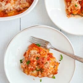 zucchini lasagna roll up on a white dinner plate with fork, dish of roll ups in background.