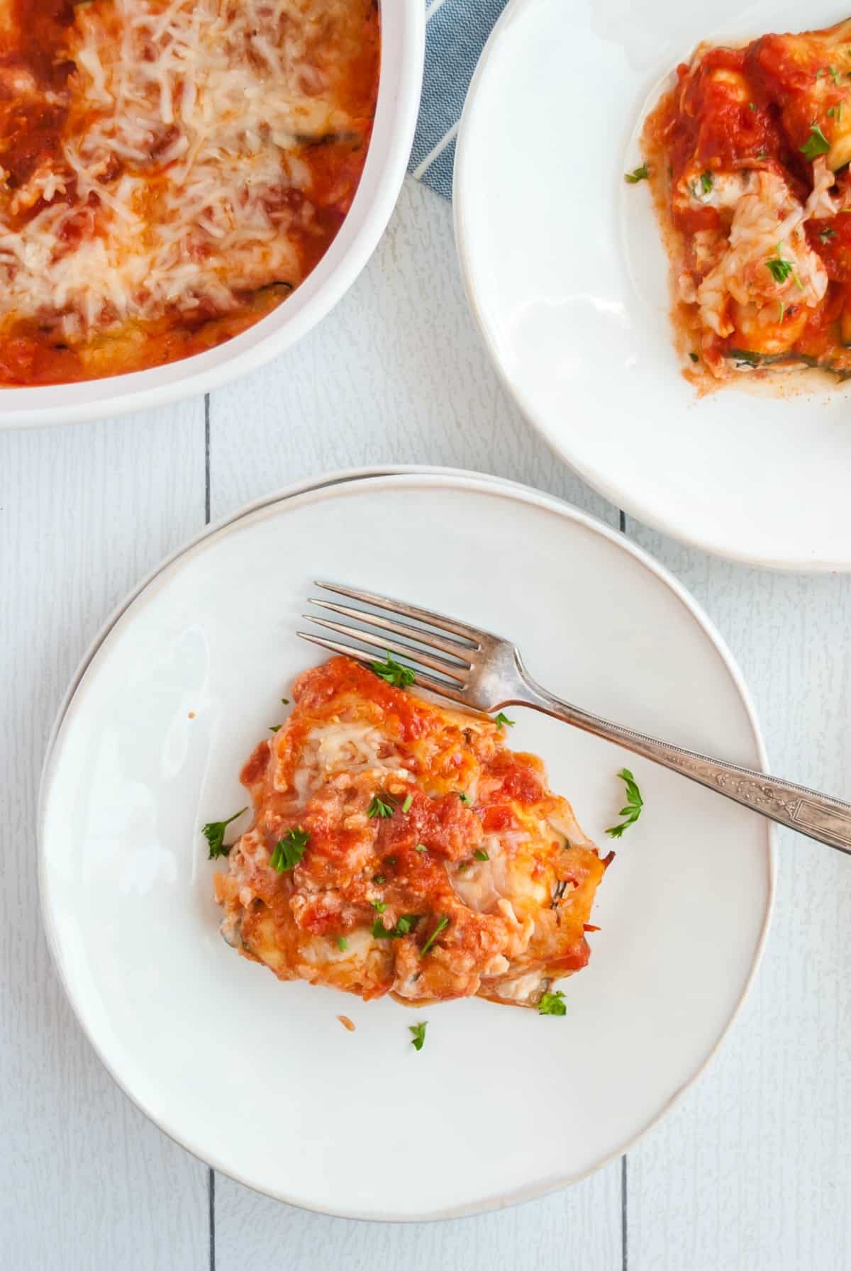 zucchini lasagna roll up on a white dinner plate with fork, dish of roll ups in background.