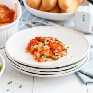 Stack of white dinner plates with zucchini lasagna roll ups on top plate. Table set with baking dish, breadsticks and salad in background.