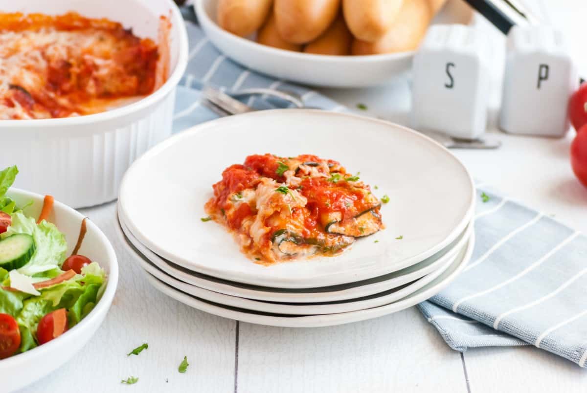 Stack of white dinner plates with zucchini lasagna roll ups on top plate. Table set with baking dish, breadsticks and salad in background.