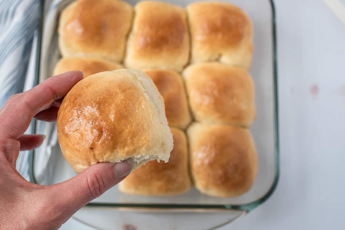 Glass square baking dish with fluffy dinner rolls, one being lifted out of the pan.