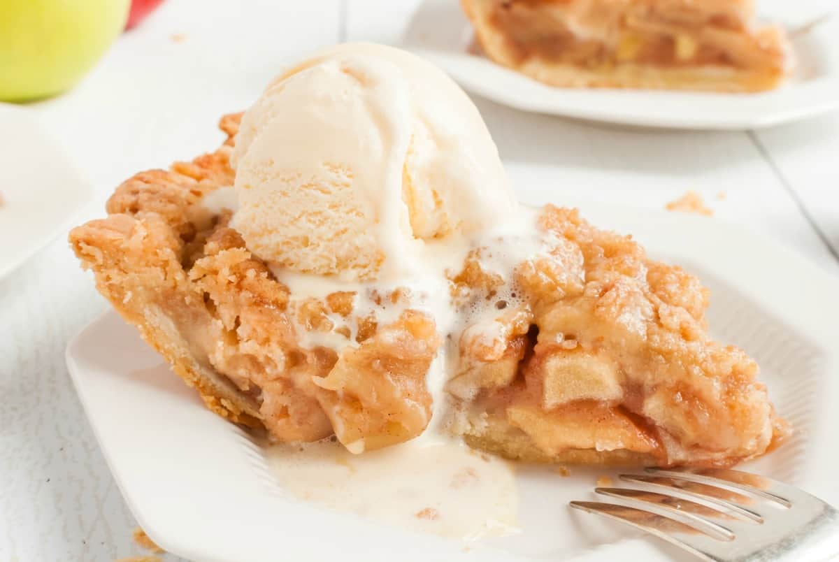 Slice of apple pie on a white dessert plate with a melting scoop of vanilla ice cream on top.