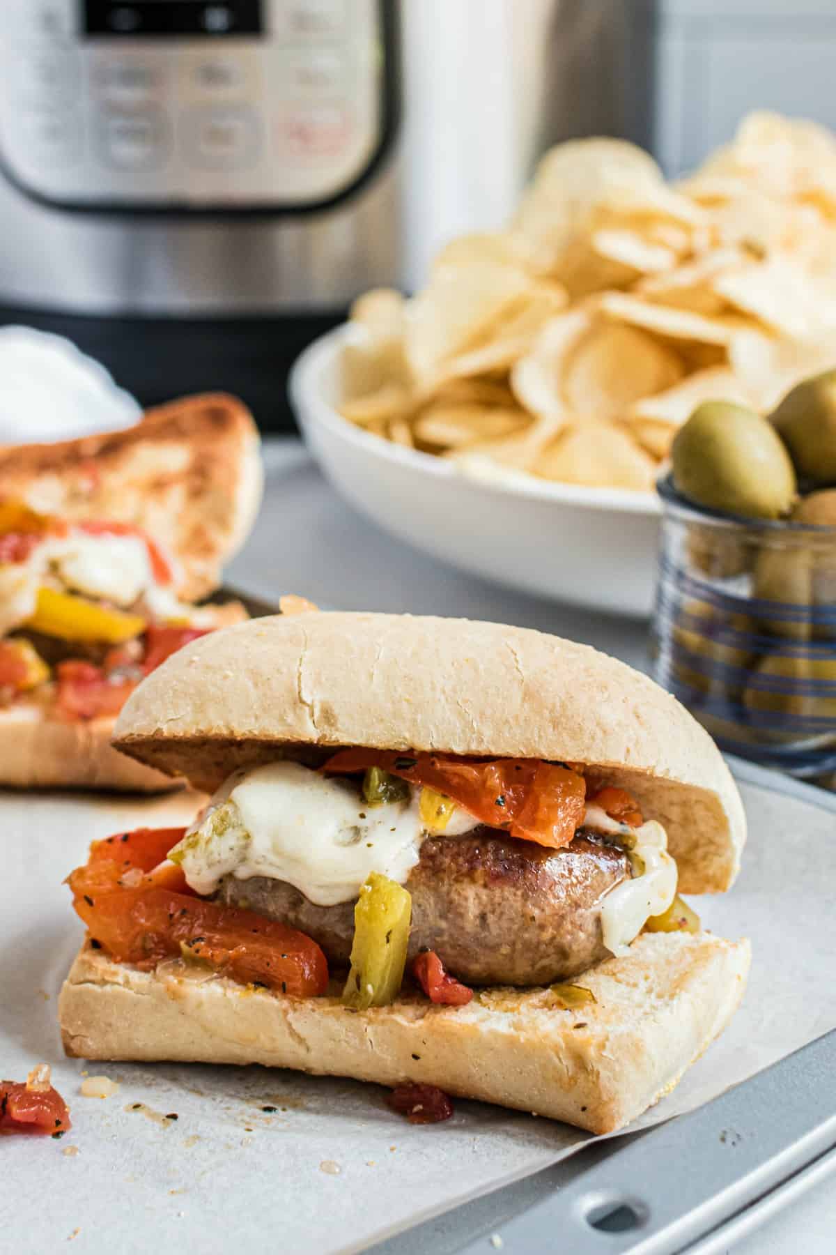 Sausage and peppers on a french roll with chips, olives, and pressure cooker in background.