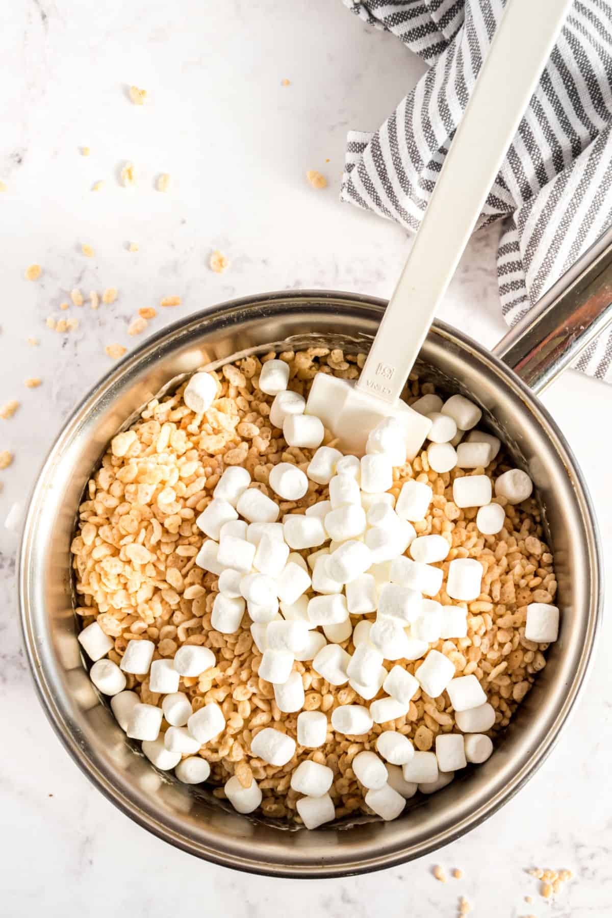 Melted butter and marshmallow for rice krispie treats in a sauce pan with cereal and more marshmallows being added.