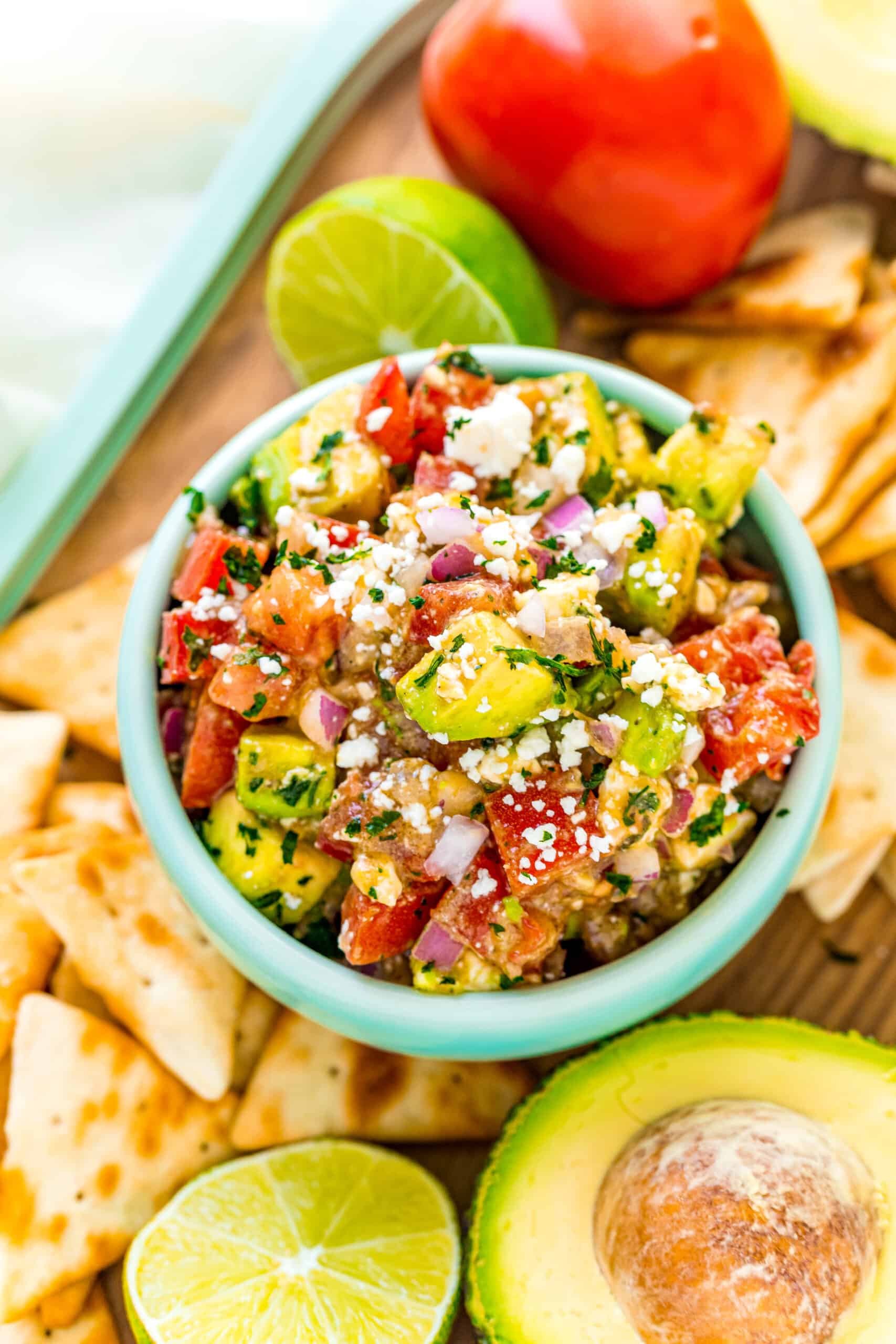 Light green bowl with avocado salsa and feta cheese, surrounded by pita chips for dipping.