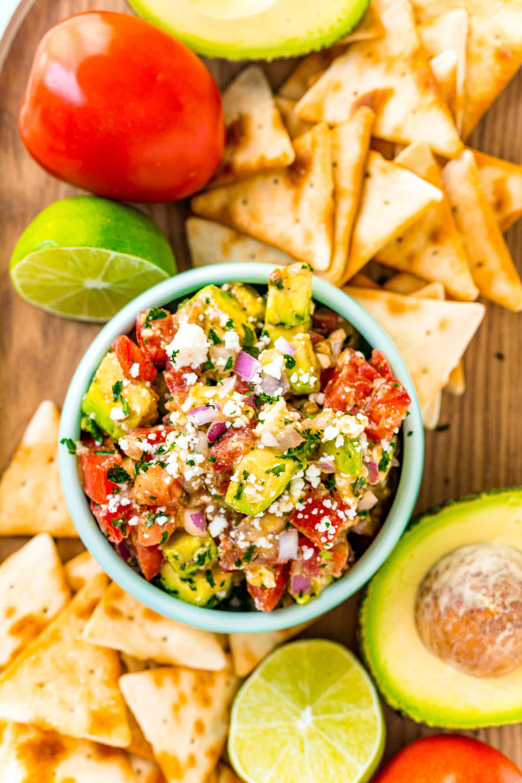 Overhead view of avocado salsa in a bowl with pita chips on a board surrounding the bowl.