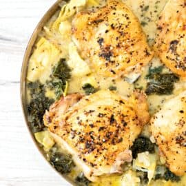 Skillet with baked chicken thighs in a creamy coconut, artichoke, and kale sauce.