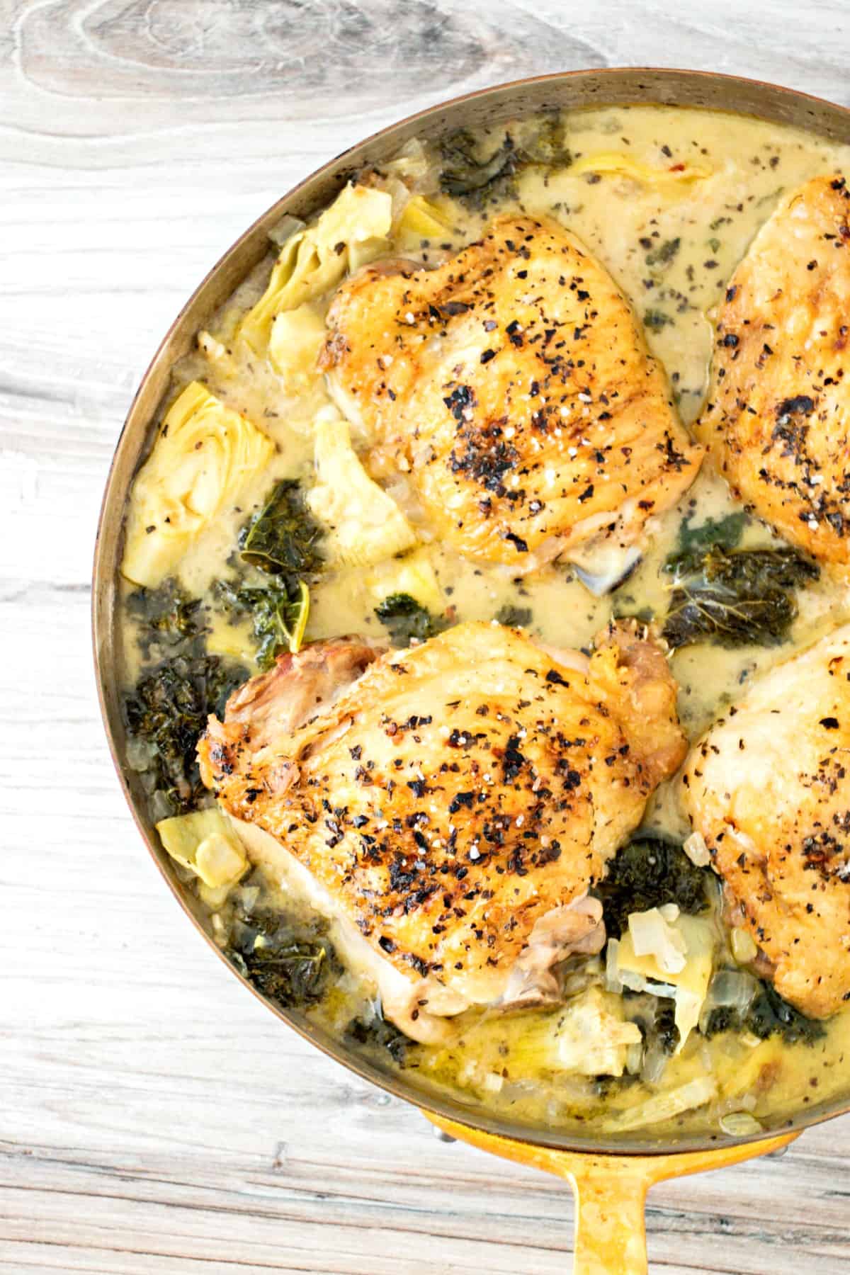 Skillet with baked chicken thighs in a creamy coconut, artichoke, and kale sauce.